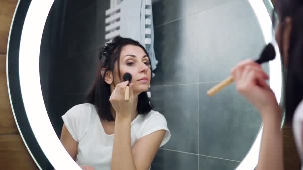 Güzel, konsantre olmuş siyah saçlı genç kadın aynaya bakıyor ve banyoda makyaj malzemesi olarak özel bir fırça kullanıyor. — Stok video