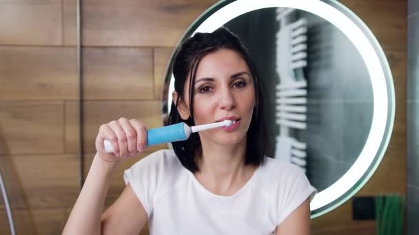 Vista frontal de la atractiva joven morena sonriente que se cepilla los dientes con pasta de dientes en el baño — Vídeos de Stock