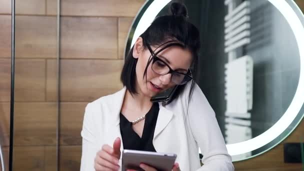 Vista da vicino della giovane donna d'affari soddisfatta di bell'aspetto con i capelli neri che indossa occhiali e vestito in abito bianco parlando al telefono e utilizzando tablet pc in bagno — Video Stock