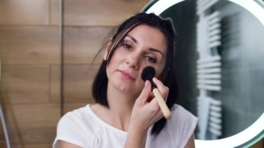 Kendine güvenen genç esmer kızın yakından görüntüsü. Fırçayla yüz pudrası uyguluyor ve kameraya bakıyor.