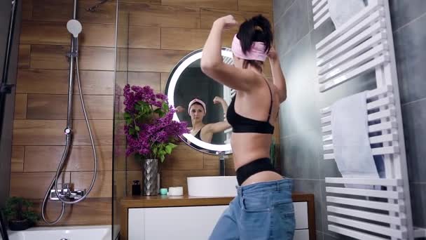 Concept de mode de vie sain où splendide jeune brune élancée en jeans de grande taille regardant son reflet dans le miroir et montrant comment elle a perdu du poids — Video