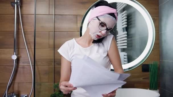 Schöne junge Frau in Kosmetikmaske telefoniert und stöbert in ihrem Badezimmer in Dokumenten — Stockvideo