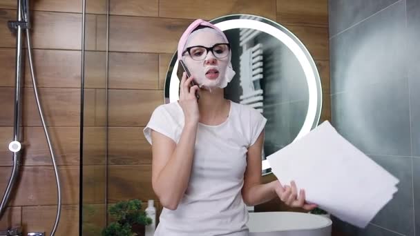 Dość emocjonalna młoda kobieta stojąca w łazience z maską kosmetyczną na twarzy i rozmawiająca przez smartfona, dająca instrukcje swojemu pracownikowi — Wideo stockowe
