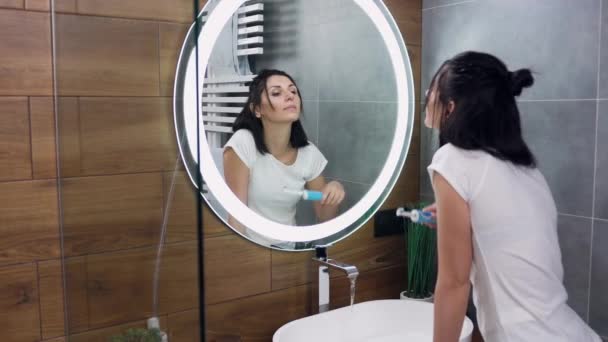 Вид збоку приємної впевненої молодої брюнетки в білій футболці, яка чистить зуби, стоїть перед дзеркалом у ванній — стокове відео
