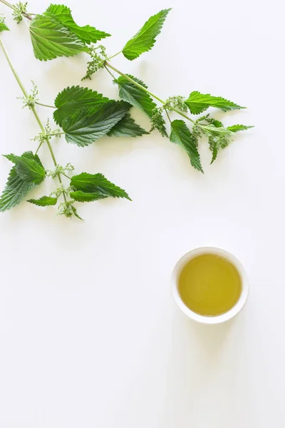 白いテーブルの上にイラクサのお茶のカップ 背景に新鮮な印象的なイラクサと 薬用植物 健康的な栄養の概念 — ストック写真