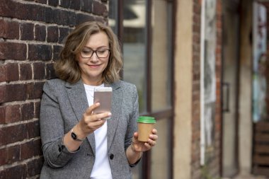 55 yaşında güzel sarışın bir kadının portresi, gülümsüyor, elinde akıllı bir telefon ve elinde kağıt bir fincan kahveyle. Açık havada, şehirde..