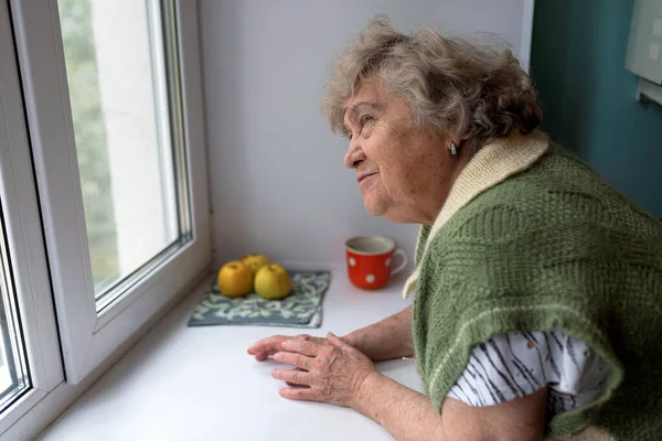 Thoughtful elderly woman at home in the kitchen looking out the window.