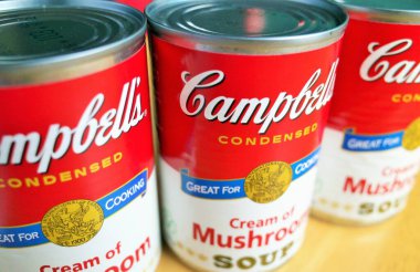 BRACKNELL, ENGLAND - FEBRUARY 05, 2020: Three tins of Campbell's Cream of Mushroom Soup on a wooden counter top with shallow focus on the center can clipart
