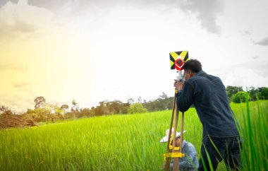Asya akıllı mühendis ya da siyah kot pantolon ve uzun kollu gömlek surveyor pirinç alan teodolit tarafından toprakları ölçme.
