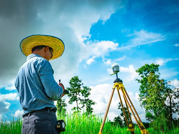 Ingeniero inteligente asiático o topógrafo en jeans negros y camisa de manga larga y sombrero de bambú tejido. Está trabajando en la pantalla del controlador para inspeccionar la tierra en el campo de arroz, Tailandia. Instrumento de topografía GPS . —  Fotos de Stock