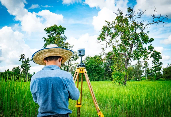 Ingénieur ou arpenteur asiatique intelligent en jeans noirs et chemise à manches longues et chapeau en bambou tissé. Il travaille sur l'écran du contrôleur pour l'arpentage des terres dans la rizière, Thaïlande. Instrument de levé GPS . — Photo