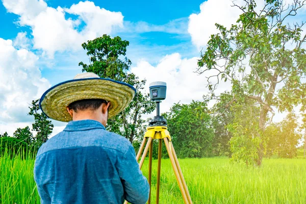 Engenheiro inteligente asiático ou topógrafo em jeans pretos e camisa de manga longa e chapéu de bambu tecido. Ele está trabalhando na tela do controlador para levantamento de terras no campo de arroz, na Tailândia. Instrumento de levantamento GPS . — Fotografia de Stock