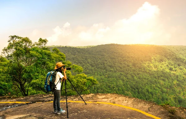 Seyahat olan kadın sırt çantası şapka ve kamera tripod stand onu tatile woods ve gökyüzü güzel manzarasını izlerken dağ uçurumun tepesinde. Asyalı kadın seyahat tek başına.