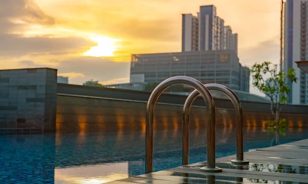 Piscina en el lujoso hotel y spa por la mañana con el cielo dorado del amanecer. Escalera de barras de agarre de primer plano con agua limpia y suelo de madera al borde de la piscina. Ocio en el hotel concepto junto a la piscina . — Foto de Stock