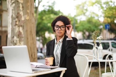 Genç Afro-Amerikan kız elinde fincan kahve ile kafeterya masada oturan gözlük ve masa üstünde laptop