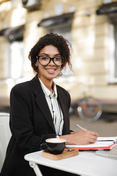 Porträt eines afrikanisch-amerikanischen Mädchens mit Brille, das am Tisch eines Cafés sitzt und glücklich mit dem Stift in der Hand in die Kamera blickt — Stockfoto