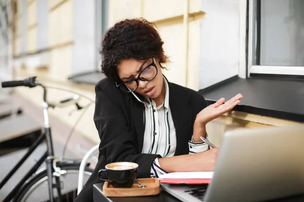 Retrato de menina afro-americana em óculos sentados à mesa e emocionalmente falando em seu celular — Fotografia de Stock