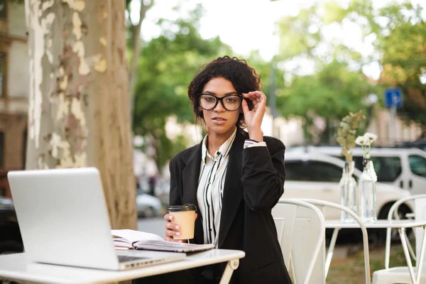 Genç Afro-Amerikan kız elinde fincan kahve ile kafeterya masada oturan gözlük ve masa üstünde laptop