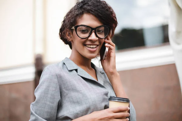 Mignonne afro-américaine fille dans des lunettes debout sur la rue et parler sur son téléphone portable avec du café dans les mains — Photo