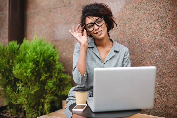 Hermosa chica afroamericana en gafas sentadas en el banco y trabajando en su computadora portátil mientras mira felizmente en la cámara —  Fotos de Stock