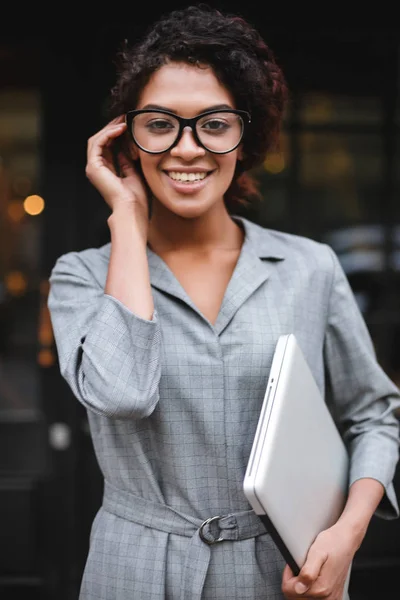Retrato de menina afro-americana sorridente em óculos de pé com laptop na mão e olhando alegremente na câmera — Fotografia de Stock