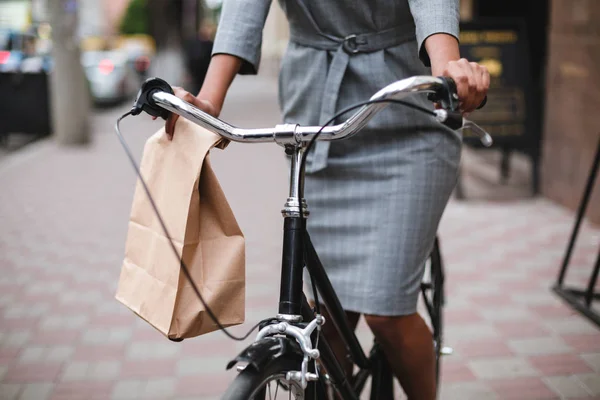 Close up photo of woman body in gray dress riding on bicycle along the street. Portrait of young lady riding bicycle with paper bag in hand — Stock Photo, Image