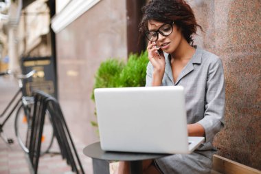 Bankta oturan ve dizüstü bilgisayarda çalışırken onu telefonda konuşurken gözlüklü Afro-Amerikan kız portresi