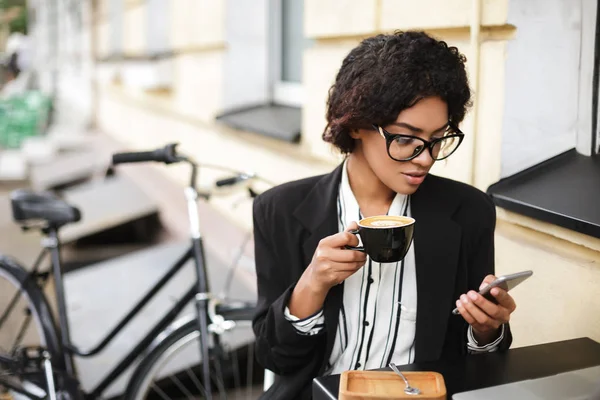 รูปภาพของสาวแอฟริกันอเมริกันในแว่นตานั่งที่โต๊ะของคาเฟ่พร้อมโทรศัพท์มือถือและถ้วยกาแฟในมือ — ภาพถ่ายสต็อก