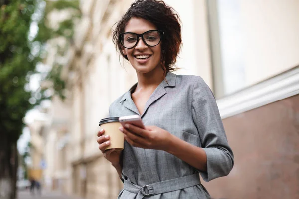 Hymyilevä afroamerikkalainen tyttö silmälaseissa seisoo kadulla kännykkä ja kahvi käsissä, kun taas onnellisesti katsot kameraan — kuvapankkivalokuva