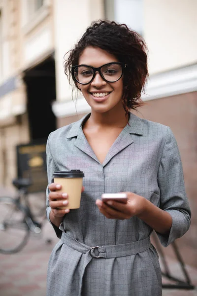 En pen afroamerikansk jente med briller som står på gata med mobiltelefon og kaffe i hendene mens hun ser med glede i kamera – stockfoto