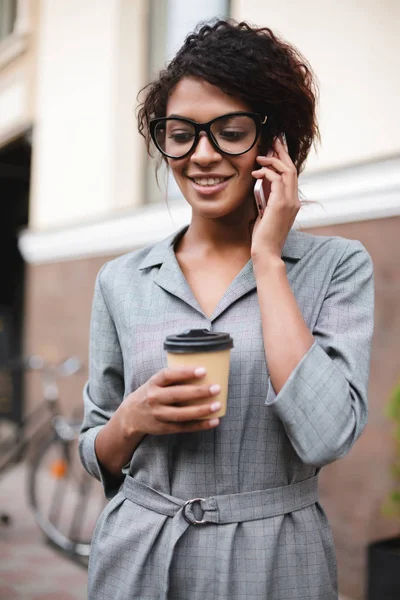 Nice africká americká dívka v brýlích stojící na ulici a mluvit na její mobil s kávou — Stock fotografie