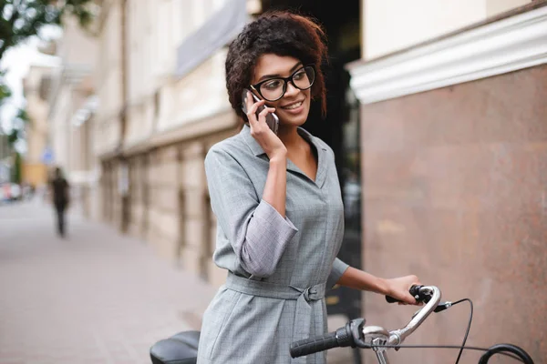 Portrett av lystig afroamerikansk jente med briller som står med sykkel og snakker på mobilen – stockfoto