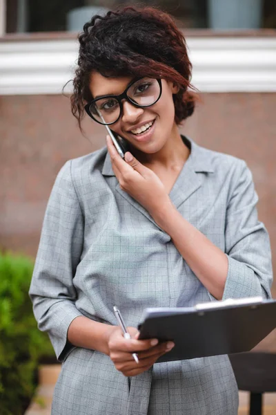 Unga leende African American flicka i glasögon står och glatt tittar i kameran medan du talar i hennes mobiltelefon — Stockfoto