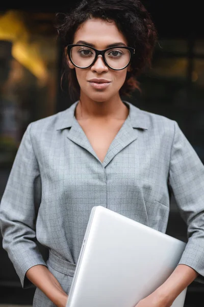 Ritratto di ragazza afroamericana pensierosa in occhiali in piedi con il computer portatile in mano e sognante guardando in macchina fotografica — Foto Stock