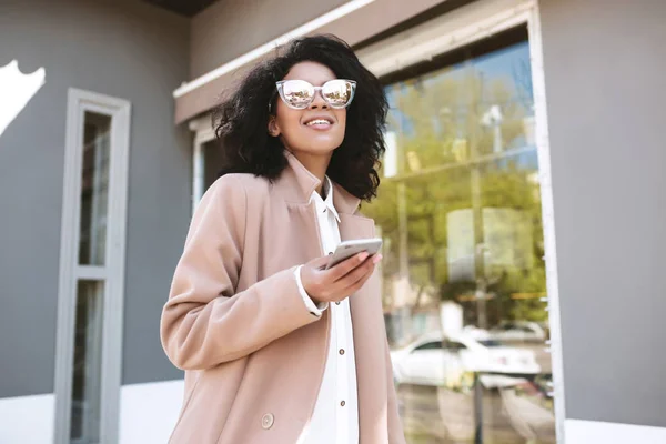 Mladá dívka v sluneční brýle na její mobil. Docela americkou afričanku v kabát, opírajíc se o plot — Stock fotografie