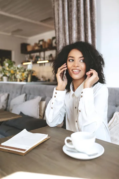 Mladá africká americká dívka sedí v restauraci a mluví na mobilním telefonu. Dívka s tmavými kudrnatými vlasy posezení v kavárně s šálkem kávy a menu na stole — Stock fotografie