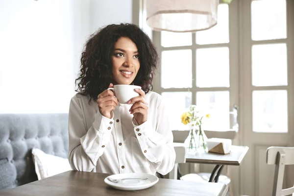 손에 커피 한잔과 함께 레스토랑에 앉아 흰 셔츠에 젊은 아프리카계 미국인 여자. 카페에서 커피를 마시는 아주 웃는 소녀 — 스톡 사진