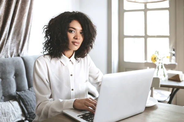 Chica joven en camisa blanca sentado en el restaurante con el ordenador portátil. Bastante chica afroamericana con pelo rizado oscuro trabajando en su computadora portátil en la cafetería — Foto de Stock