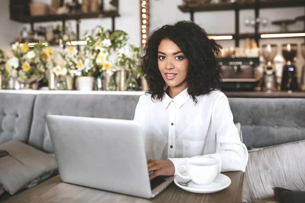 Joven chica afroamericana sentada en el restaurante con portátil y taza de café en la mesa —  Fotos de Stock