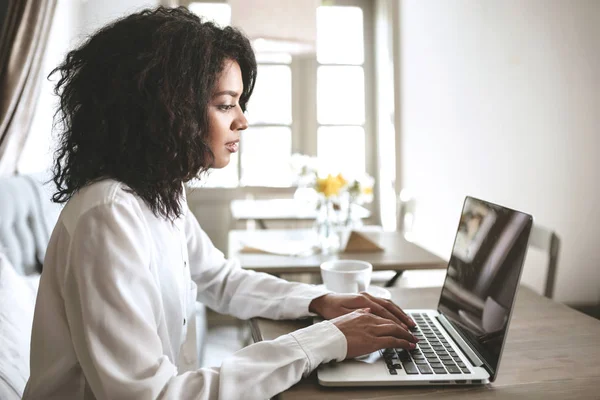 Joven chica afroamericana trabajando en su computadora portátil en restaurante.Pretty dama con el pelo rizado oscuro sentado en la cafetería con computadora portátil —  Fotos de Stock