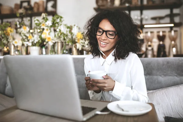Νέοι Αφρικανικός Αμερικανός κορίτσι με τα γυαλιά, κάθεται στο εστιατόριο με φορητό υπολογιστή και το φλιτζάνι του καφέ στα χέρια — Φωτογραφία Αρχείου