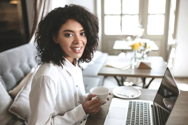 Chica sonriente joven mirando en la cámara con la taza en las manos en la café.Chica afroamericana bastante sentado en el restaurante con el ordenador portátil —  Fotos de Stock