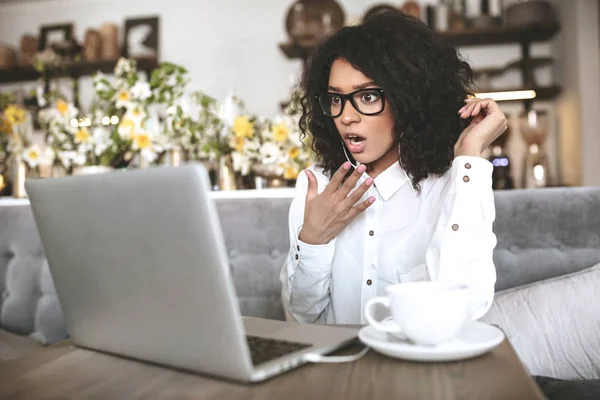 Chica joven en gafas sorprendentemente mirando en el ordenador portátil en la café. chica afroamericana sentada en el restaurante con el ordenador portátil y la taza de café en la mesa — Foto de Stock