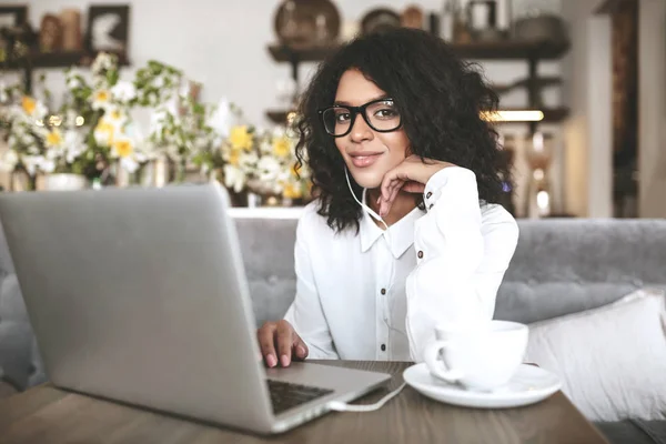 Niza chica afroamericana en gafas sentado en el restaurante con el ordenador portátil y la taza de café.Joven con el pelo rizado oscuro trabajando en el ordenador portátil en los auriculares —  Fotos de Stock