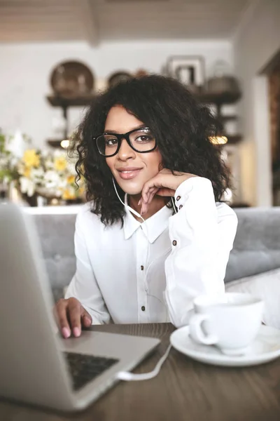 Chica afroamericana bonita en gafas sentado en el restaurante con el ordenador portátil y la taza de café.Joven con el pelo rizado oscuro trabajando en el ordenador portátil en los auriculares —  Fotos de Stock