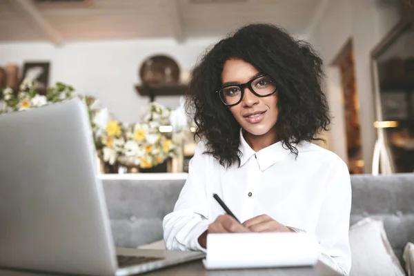 Niza chica afroamericana en gafas de trabajo en el restaurante con ordenador portátil y notebook.Young dama con pelo rizado oscuro sentado en la cafetería —  Fotos de Stock