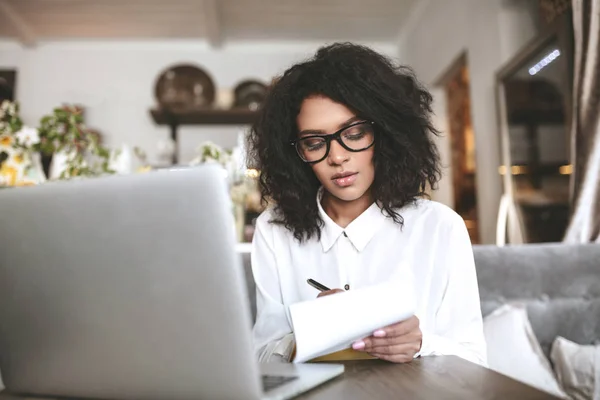 Junge afrikanisch-amerikanische Mädchen mit Brille sitzt im Restaurant und schreibt Notizen. Hübsches Mädchen mit dunklem lockigem Haar arbeitet im Café mit Laptop — Stockfoto