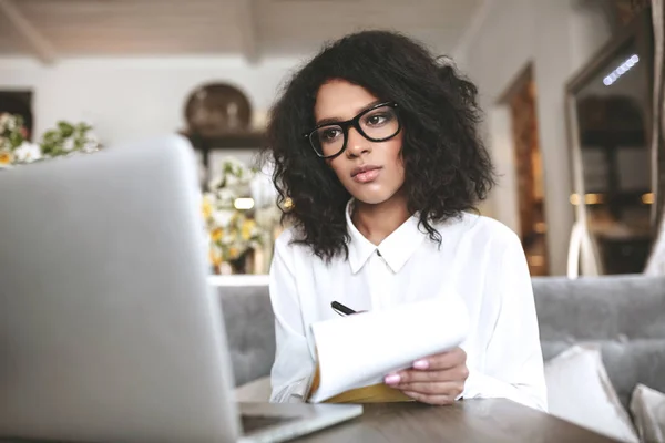 Chica afroamericana bonita en gafas de trabajo en el ordenador portátil en restaurante.Joven con el pelo rizado oscuro sentado en la cafetería con portátil en la mano —  Fotos de Stock