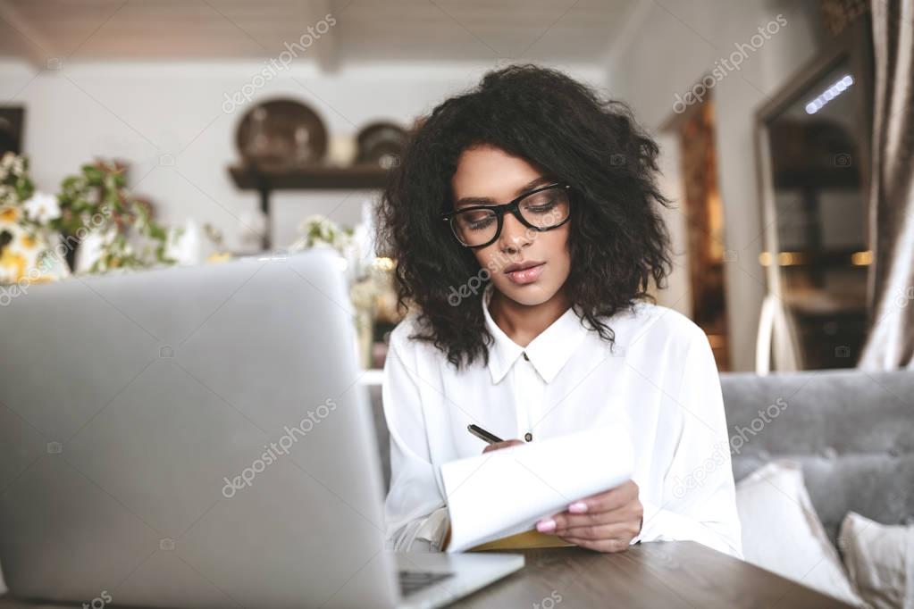 Young African American girl with glasses sitting in restaurant and writing notes.Pretty girl with dark curly hair working in cafe with laptop