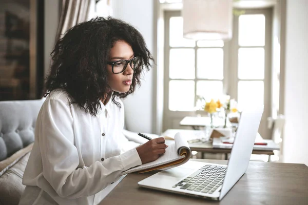 Chica afroamericana joven escribiendo notas en restaurante.Nice chica con pelo rizado oscuro sentado en la cafetería con ordenador portátil y portátil — Foto de Stock